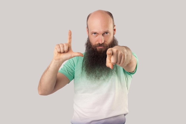 Foto sos un perdedor. retrato de un hombre calvo barbudo de mediana edad con una camiseta verde de pie con un gesto perdedor y señalando, mirando la cámara regañando. tiro de estudio interior, aislado sobre fondo gris.