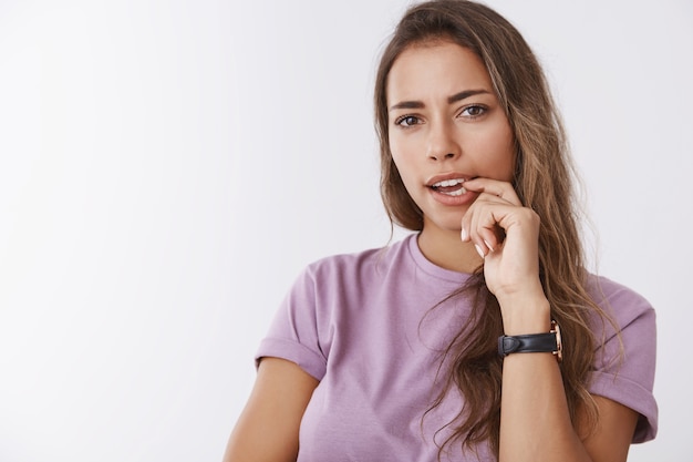 Sospechosa, decidida y atractiva mujer pensando de pie pensativo tiene dudas mordiendo el dedo entrecerrar los ojos inseguro, juzgando, tomando una decisión, dudando de pie pared blanca interesada