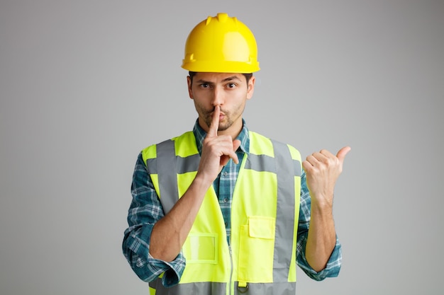 Sospechoso joven ingeniero masculino con casco de seguridad y uniforme mirando a la cámara mostrando un gesto silencioso apuntando al lado aislado en fondo blanco