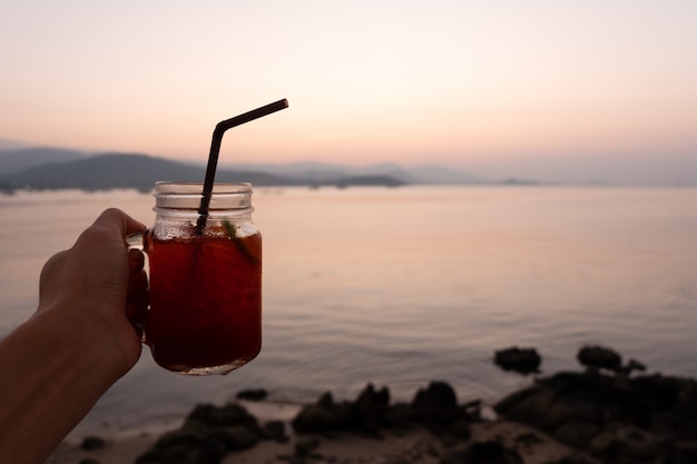 Foto dé sostener el vidrio de té de hielo del limón en la playa tropical del mar en puesta del sol.