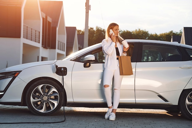 Sosteniendo una bolsa de compras Mujer joven vestida de blanco está con su auto eléctrico durante el día