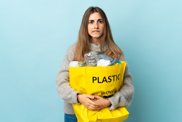 Sosteniendo una bolsa llena de botellas de plástico para reciclar sobre azul aislado con dudas y con expresión de la cara confusa