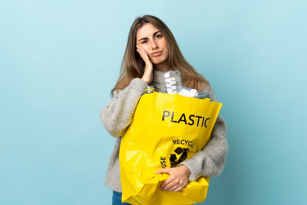 Sosteniendo una bolsa llena de botellas de plástico para reciclar sobre azul aislado infeliz y frustrado