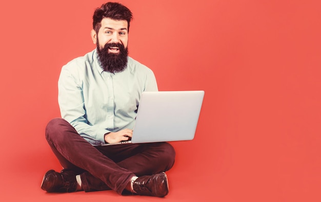 Sosteniendo la computadora portátil. Sonriente portátil trabajador guapo barbudo. Feliz joven sentado en el suelo con y usando la computadora portátil sobre fondo rojo.