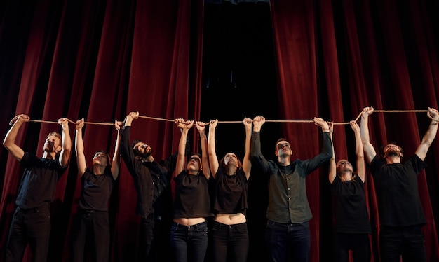 Sosteniendo la cuerda en las manos por encima de la cabeza Grupo de actores en ropa de color oscuro en el ensayo en el teatro