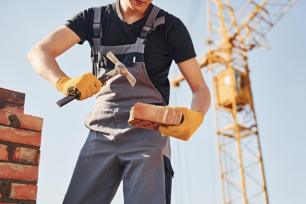 Sosteniendo ladrillos y usando martillo Trabajador de la construcción en uniforme y equipo de seguridad tiene trabajo en la construcción