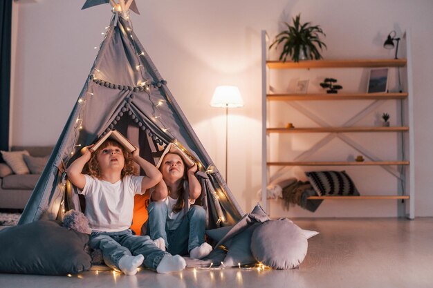 Sosteniendo libros en la cabeza Estado de ánimo juguetón Dos niñas pequeñas están juntas en la carpa en la habitación doméstica