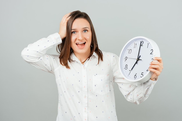 Foto sosteniendo la mano en la cabeza, una mujer joven sostiene un reloj de pared redondo blanco foto de estudio sobre fondo gris