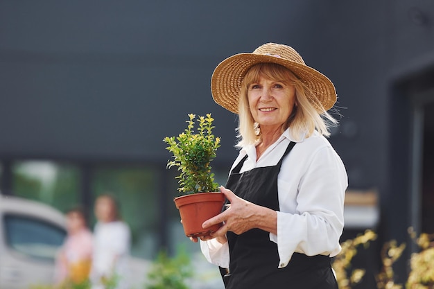 Sosteniendo la olla en las manos La mujer mayor está en el mini jardín durante el día Exterior del edificio detrás