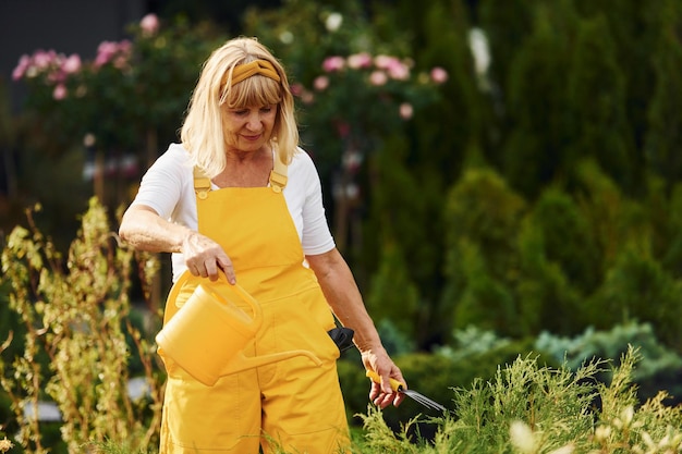 Sosteniendo una regadera amarilla La mujer mayor está en el jardín durante el día Concepción de las plantas y las estaciones