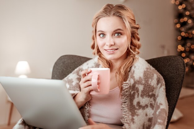 Sosteniendo la taza llena. Hermosa freelance de pelo largo sentada en un cómodo sillón mientras está sola en sus apartamentos