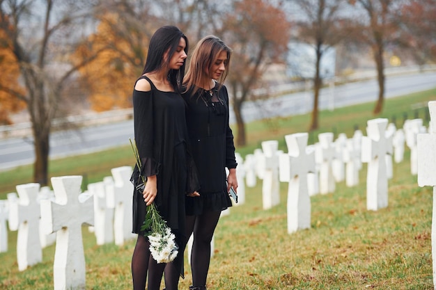 Sostiene flores Dos mujeres jóvenes vestidas de negro visitando el cementerio con muchas cruces blancas Concepción del funeral y la muerte