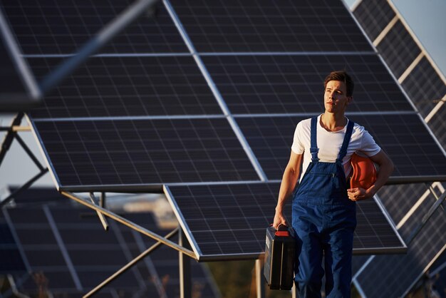 Sostiene el maletín con el equipo en la mano. Trabajador de sexo masculino en uniforme azul al aire libre con baterías solares en un día soleado.