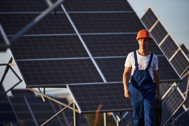 Sostiene el maletín con el equipo en la mano. Trabajador de sexo masculino en uniforme azul al aire libre con baterías solares en un día soleado.