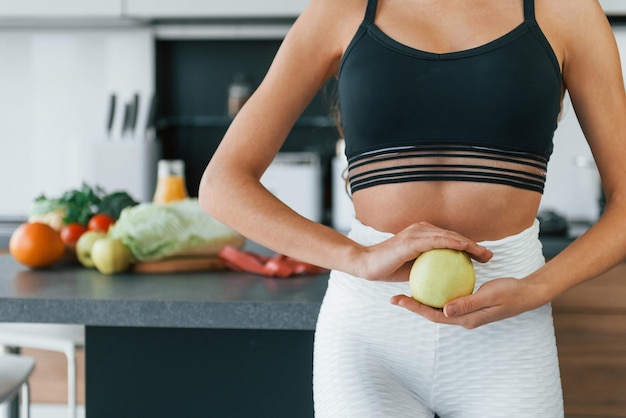 Sostiene la manzana contra el vientre La joven europea está en el interior de la cocina con comida saludable