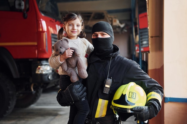 Sostiene osito de peluche La niña feliz está con un bombero en uniforme protector