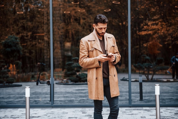 Sostiene el teléfono inteligente Modelo masculino joven en ropa de moda está al aire libre en la ciudad durante el día