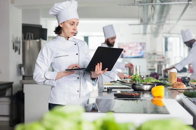 Sous chef com laptop moderno procurando receita de prato gourmet em pé na cozinha profissional do restaurante. Trabalhador da indústria de alimentos com idéias de enfeite de brainstorming de computador portátil.