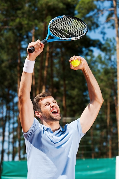 ¡Soy un ganador! Feliz joven en camisa de polo levantando su raqueta de tenis mientras está de pie en la cancha de tenis