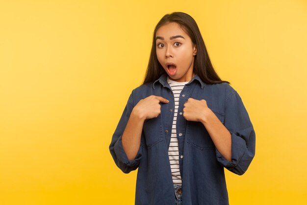 Este soy yo Retrato de una chica sorprendida con camisa de mezclilla apuntándose a sí misma y mirando con expresión de asombro gritando de una repentina e increíble noticia loca en un estudio interior aislado en un fondo amarillo