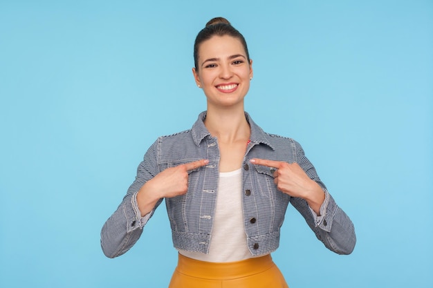 Este soy yo Retrato de una mujer feliz, segura de sí misma, vestida a la moda, con un moño en el pelo, apuntándose a sí misma y sonriendo alegremente, sintiéndose orgullosa y exitosa, en un estudio interior aislado en un fondo azul