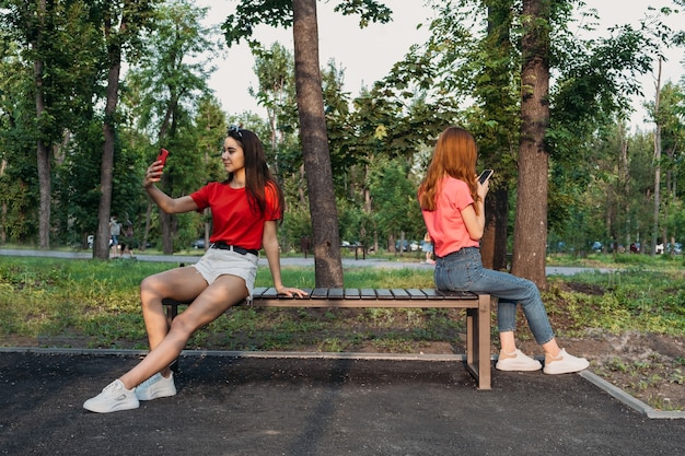 Soziale Distanzierung, neues normales Konzept. Zwei Freundinnen mit Handy sitzen auf Bank