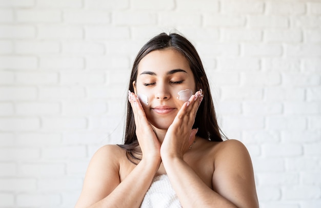 Spa y belleza. Feliz hermosa mujer caucásica vistiendo batas de baño aplicando crema facial en su rostro