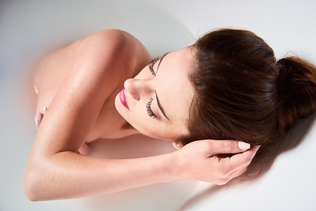 Spa em casa - a mulher relaxa no banheiro. Linda mulher caucasiana na banheira com leite. Uma garota atraente relaxante no banho na luz de fundo