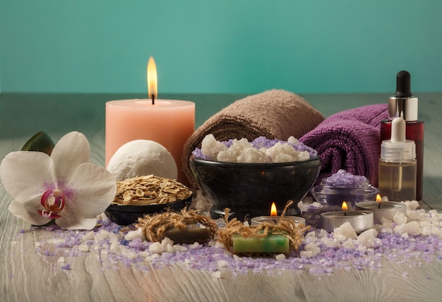 Spa-Komposition mit Orchideenblüte, Schüssel mit Meersalz, Flaschen mit Aromaöl, Seife, Peeling, Kerzen und Handtüchern auf Holzbrett und hellgrünem Hintergrund