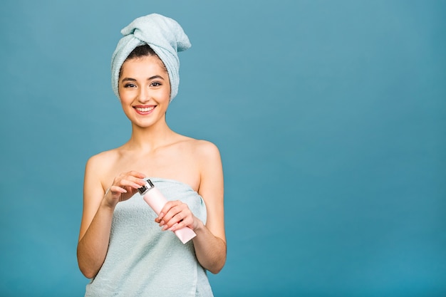 Spa-Zeit! Porträt der lächelnden schönen Frau im Handtuch über Körper und auf ihrem Kopf, der Körperlotion lokalisiert über blauem Hintergrund hält.