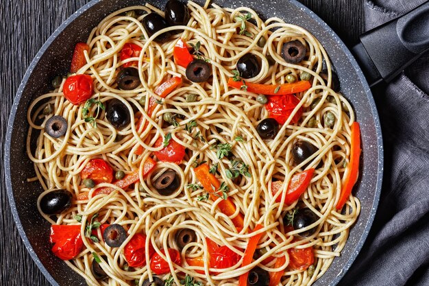 Spaghetti alla caponata de pasta integral con tomates, aceitunas y alcaparras en una sartén sobre una mesa de madera oscura, macro