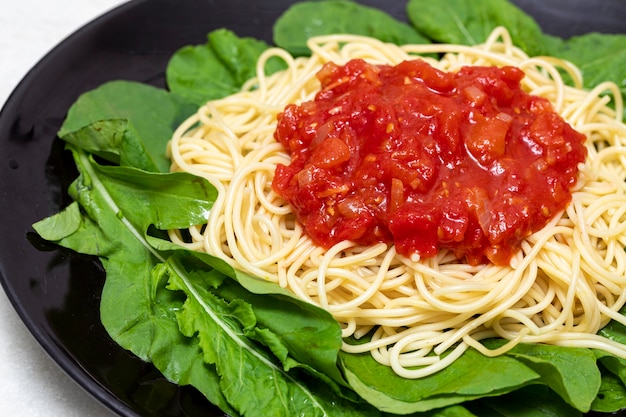 Spaghetti-Nudeln mit roter Sauce und Rucola auf schwarzem Teller mit weißem Marmorhintergrund