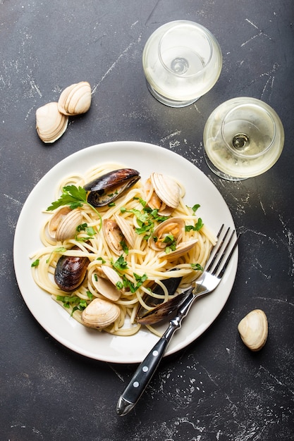 Spaghetti vongole, macarrão italiano de frutos do mar com amêijoas e mexilhões, em prato com ervas e dois copos de vinho branco sobre fundo de pedra rústico.