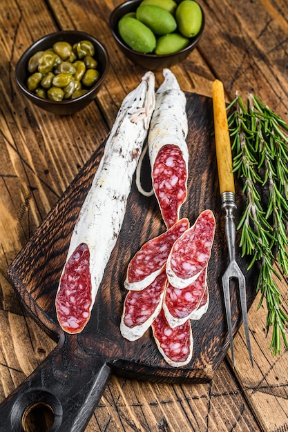 Spanische Tapas Fuet Salami Wurstscheiben mit Oliven und Rosmarin auf einem Holzbrett auf Holztisch. Draufsicht.