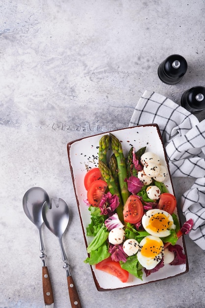 Sparagus Tomatensalat Mozzarella schwarzer Sesam Flachsöl Olivensalat und weich gekochtes Ei auf rechteckiger Keramikplatte auf grauem Betontisch Hintergrund Gesundes Ernährungskonzept Draufsicht