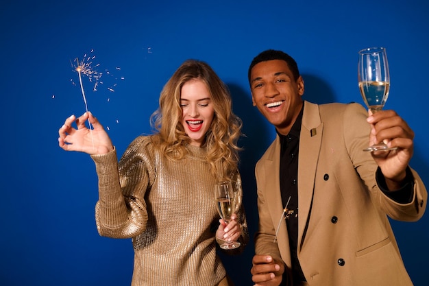 Sparklers. Menina bonita feliz com longos cachos loiros e cara de pele escura, terno com champanhe e estrelinhas queimando, comemorando sobre fundo azul