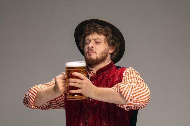 Spaß. Glücklicher Mann in traditioneller österreichischer oder bayerischer Tracht, der mit einem Krug Bier auf grauem Studiohintergrund gestikuliert. Exemplar. Die Feier, Oktoberfest, Festival, Traditionskonzept.