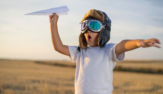 Spaß Kleiner Junge trägt Helm und träumt davon, Flieger zu werden, während er bei Sonnenuntergang ein Papierflugzeug spielt