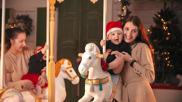 Spaß mit Weihnachtsdekoration Zwei lächelnde Mütter, die mit ihren Babys in der Nähe des Karussells stehen