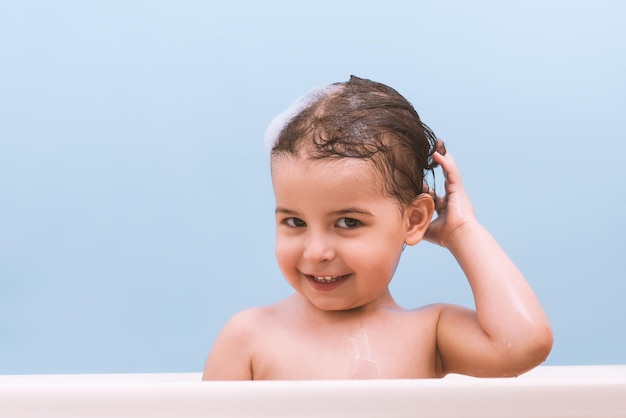 Spaß niedlich glücklich Kleinkind Baby ein Bad spielen mit Schaumblasen und Haare waschen Kleines Kind in einer Badewanne Lächelndes Kind im Badezimmer auf blauem Hintergrund Säuglingsbaden Hygiene und Gesundheitsfürsorge