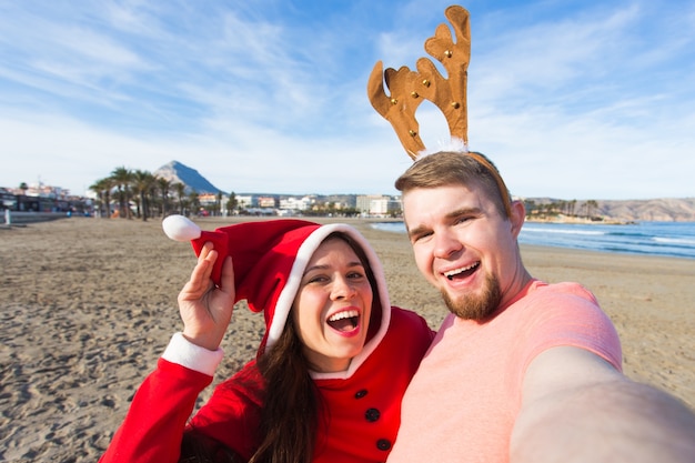 Spaß- und Winterferienkonzept - glückliches Paar in den Weihnachtskostümen, die selfie über Sandstrand nehmen