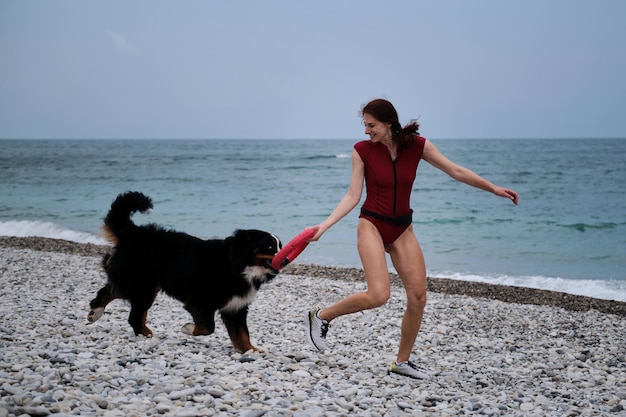 Spaziergang mit großem Hund an warmen Sommermorgen Kaukasische hübsche rothaarige Frau in Turnschuhen und Berner Sennenhund mit rotem Spielzeugring im Mund laufen schnell und spielen aktiv am Kiesstrand