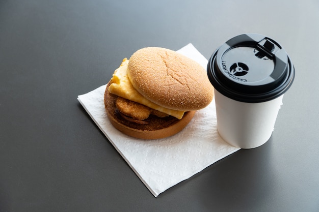 Speck- und Omelett-Hamburger auf dem Papier mit Weißbuchkaffeetasse auf dem Tisch im Schnellrestaurant