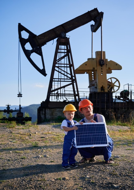 Spezialist für die Ölproduktionsindustrie mit kleinem männlichen Kind, das eine Solarbatterie in den Händen gegen eine Stabpumpe in der Ölkompressorstation hält. Alternatives Ökoenergiekonzept