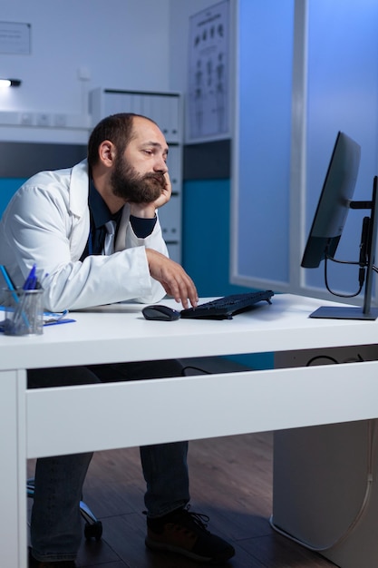 Spezialist schläft ein, während er am Schreibtisch am Computer arbeitet. Erschöpfter Arzt mit Kopf in der Hand, der nach Stunden auf den Monitor für die Gesundheitsarbeit schaut. Sanitäter arbeitet spät mit dem Gerät