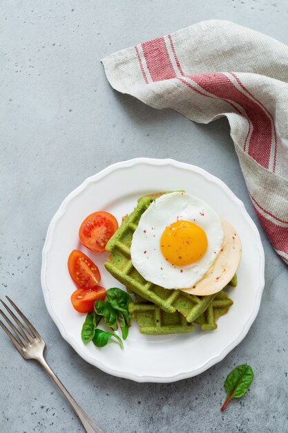 Spiegelei mit Käse, Tomaten, Mangold und Waffel mit Spinat auf weißer Keramikplatte auf hellgrauem Betontisch. Sandwich zum Frühstück. Selektiver Fokus. Draufsicht. Speicherplatz kopieren.