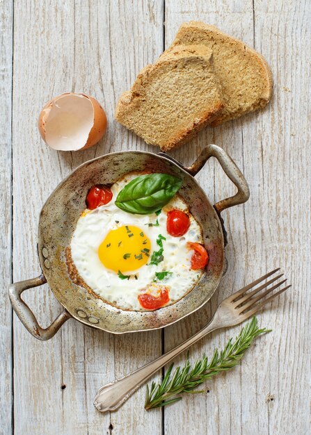 Spiegelei mit Tomaten, hausgemachtem Brot und Kräutern auf einer alten Pfanne auf Holz Draufsicht