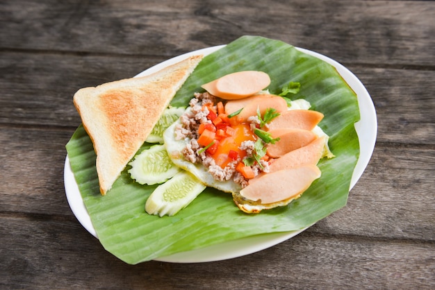 Spiegeleier mit Wurstgurkenschweinefleisch und Brot zum Frühstück auf Bananenblatthintergrund