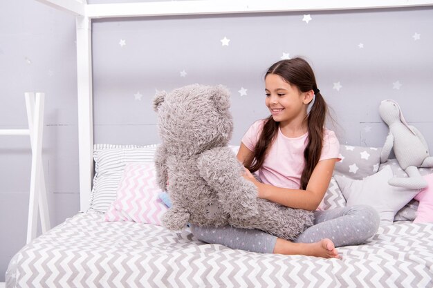 Spiele spielen Lieblingsspielzeug Mädchen Kind Umarmung Teddybär in ihrem Schlafzimmer Angenehme Zeit im gemütlichen Schlafzimmer Mädchen Kind lange Haare niedlichen Schlafanzug entspannen und spielen Plüsch-Teddybär-Spielzeug Imaginäre Interaktion