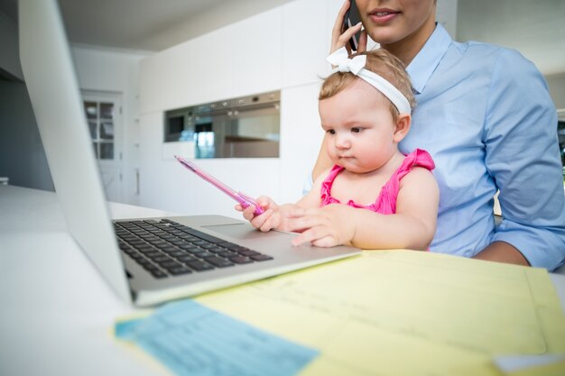 Spielerisches Baby, das mit Mutter durch Laptop sitzt